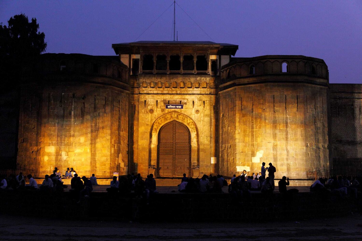 Nighttime view of a lit stone fortress gate with an arched central doorway and people gathered in front.