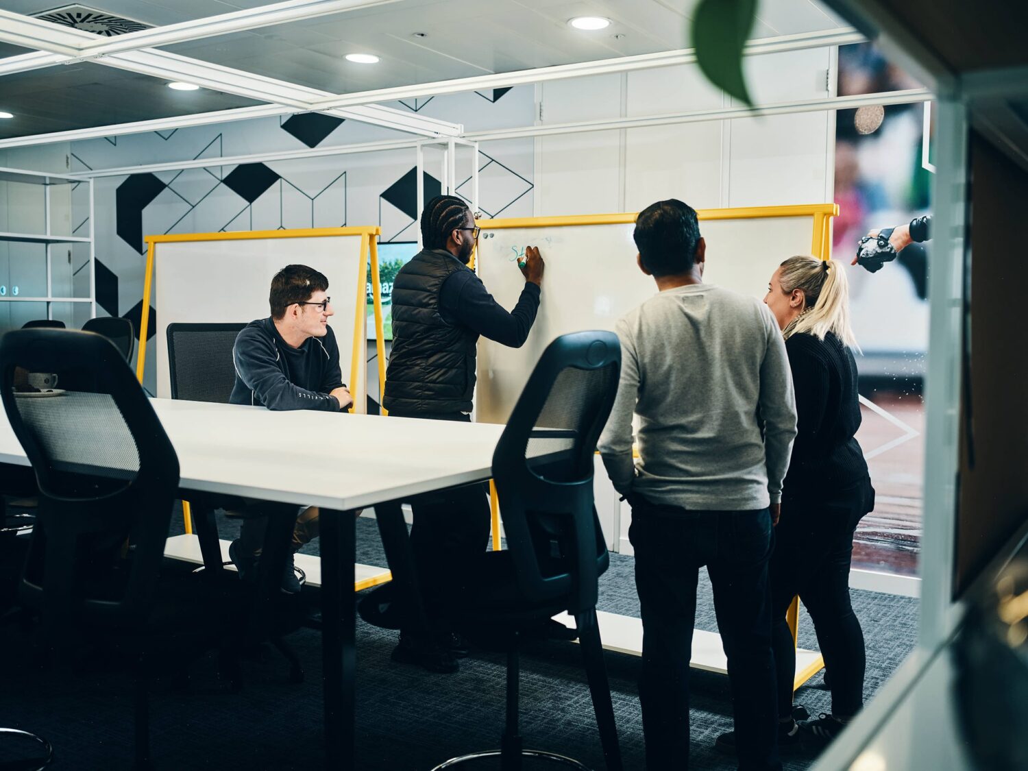 A group of people collaborates around a white table in a bright modern office, with some standing and gesturing toward a large yellow-framed whiteboard.