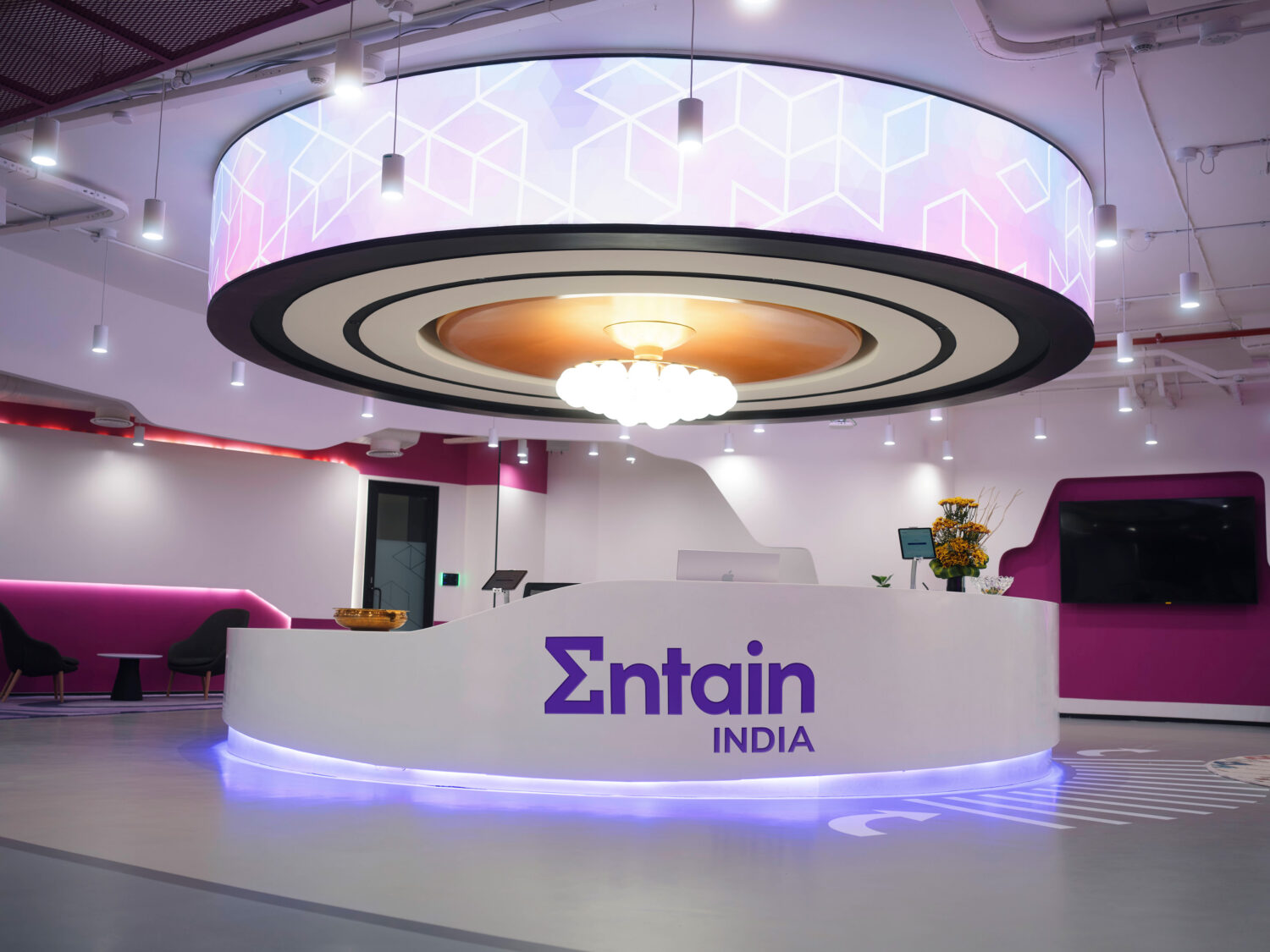 A modern exhibition booth with a white curved reception desk labeled “Entain India,” purple accent lighting, and a large circular hanging ceiling light.
