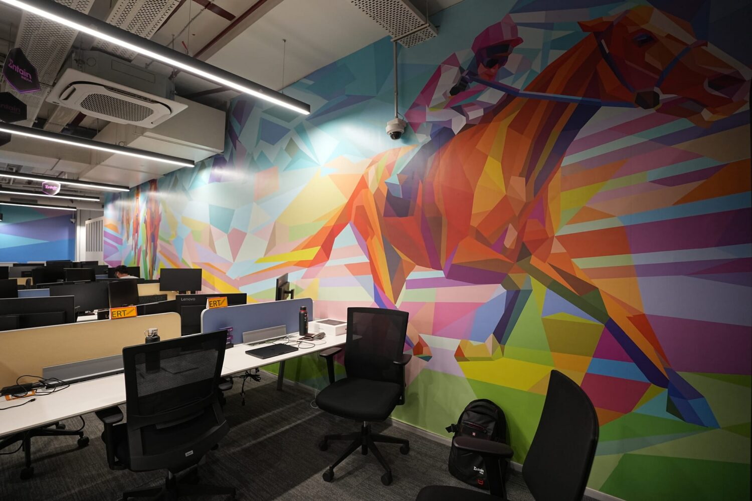 Open-plan office space with rows of desks and black chairs beside a vivid geometric mural on the wall.