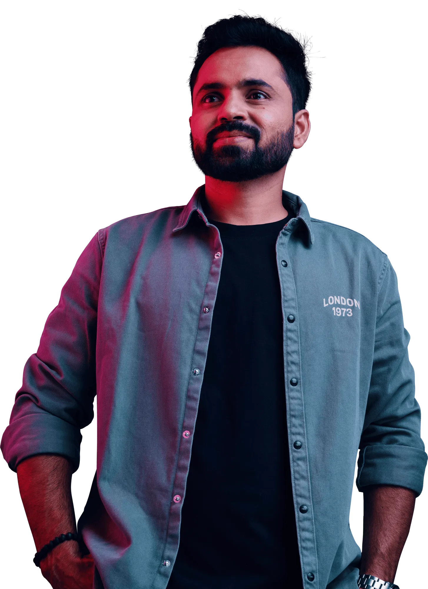 A bearded man in a light blue denim shirt over a black t-shirt stands with his hands in his pockets against a dark background.