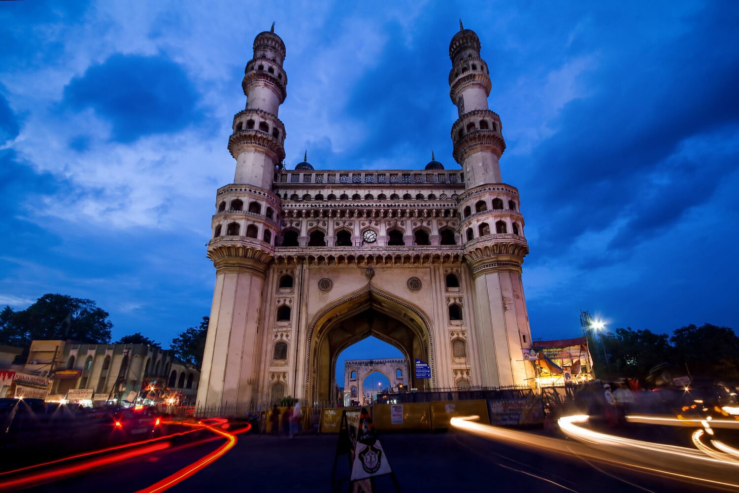 Charminar monument at night, illuminated against a blue sky with light trails from passing cars.
