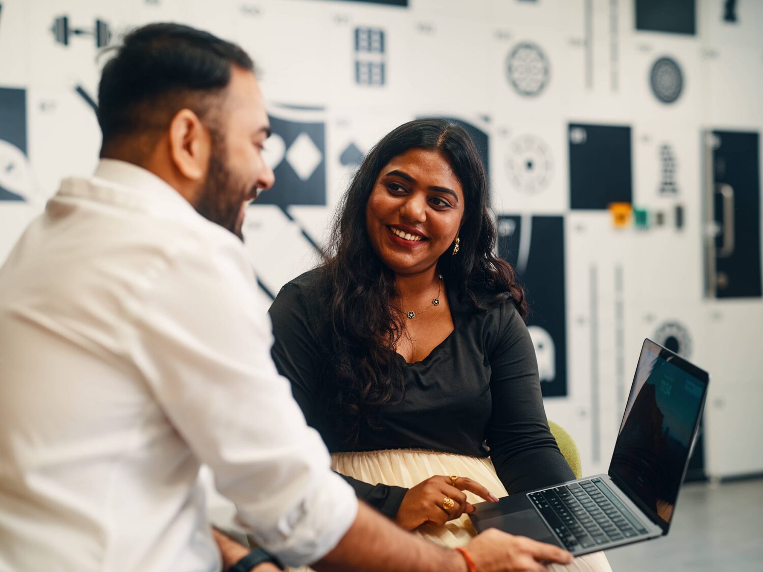 A man with a beard and a woman sit at a desk in a bright office, smiling at a laptop as they discuss.