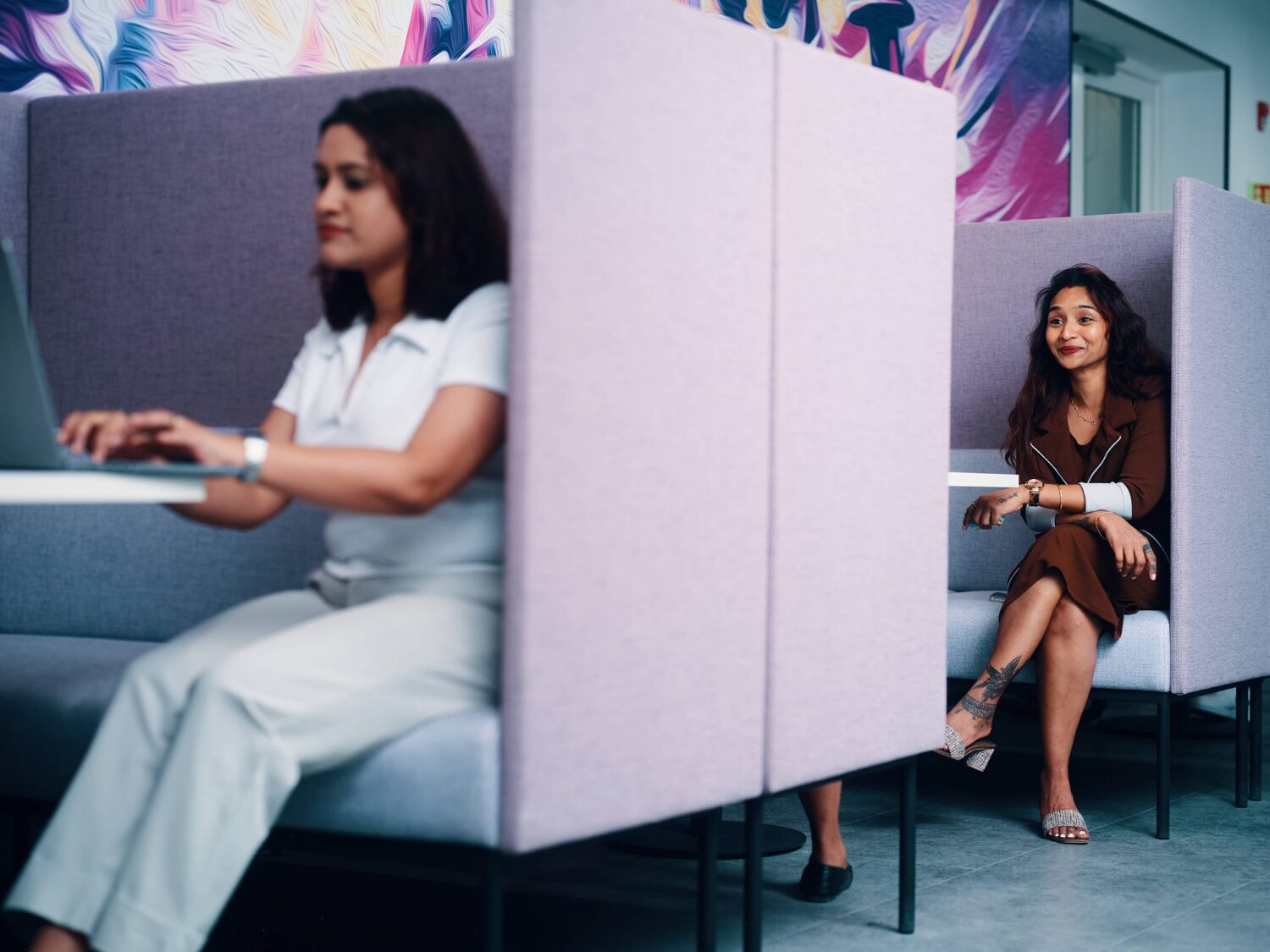 Two women sit in individual purple booths at a modern workspace, one using a laptop and the other reading a notebook.