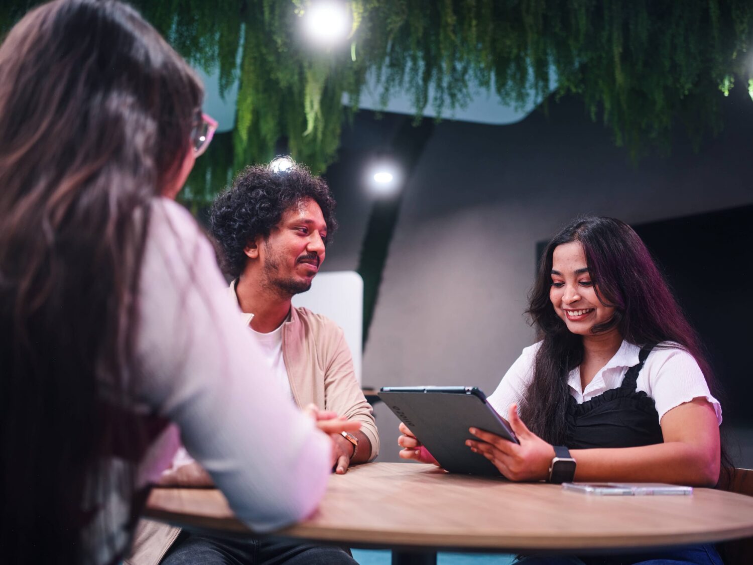 Three people sit around a round wooden table in a modern space, smiling as one person shows a tablet.