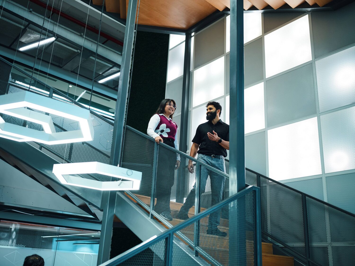Two people stand on a glass-railed mezzanine in a modern office interior with large windows and geometric pendant lights.