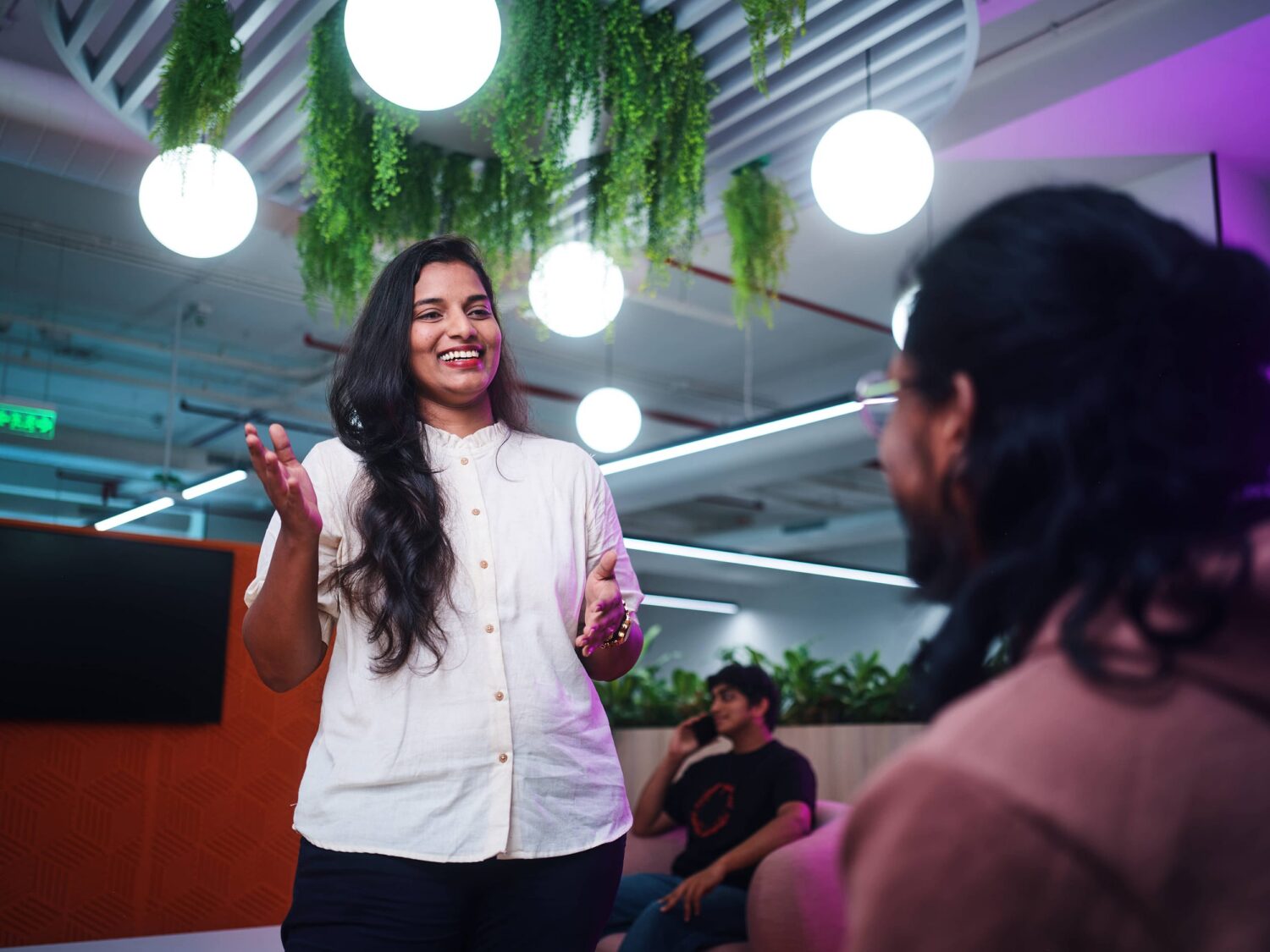 A woman in a white blouse speaks to an audience in a modern room with hanging plants and round ceiling lights.