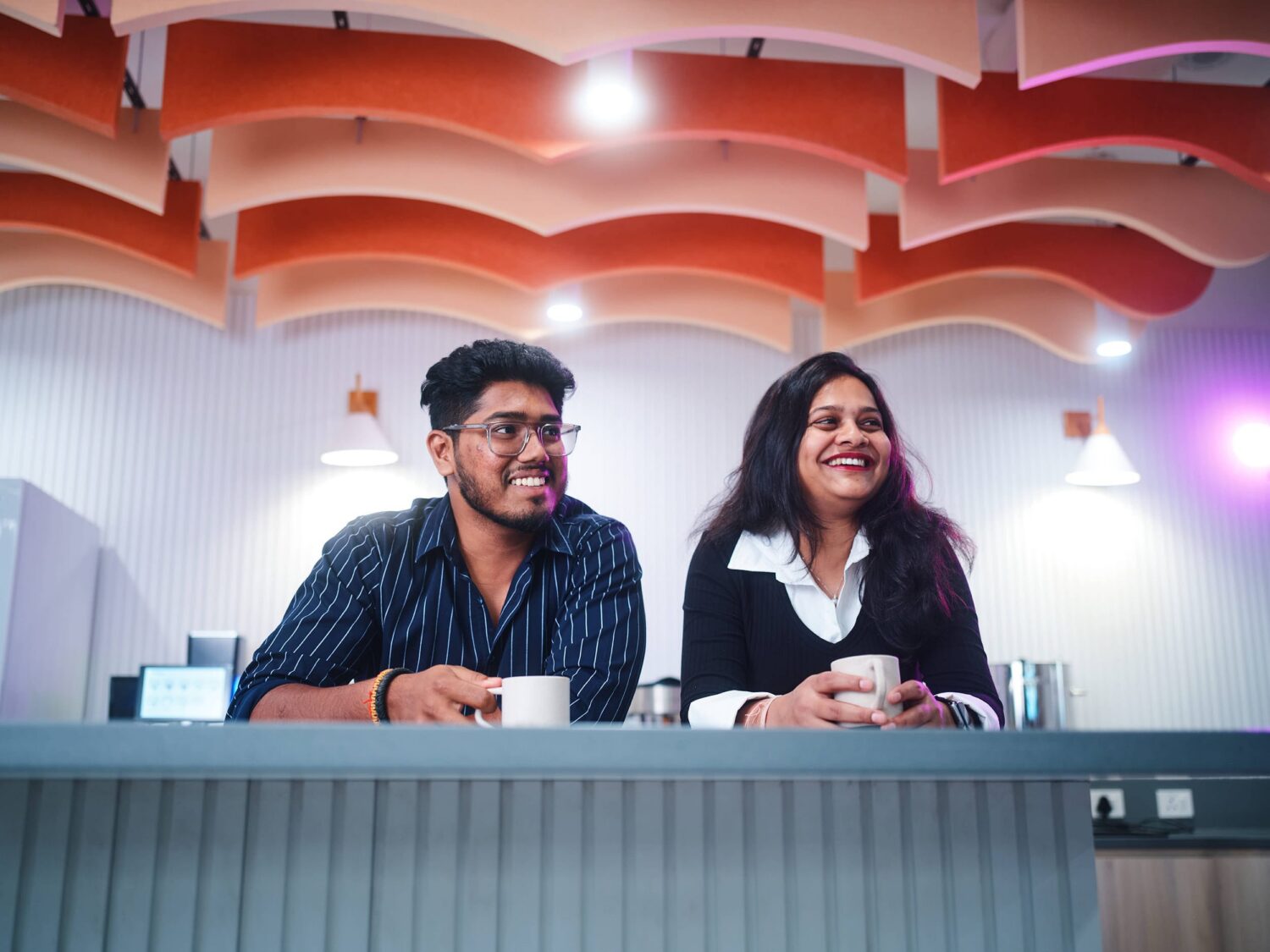 A man and a woman sit side by side behind a counter in a bright, modern cafe, smiling.