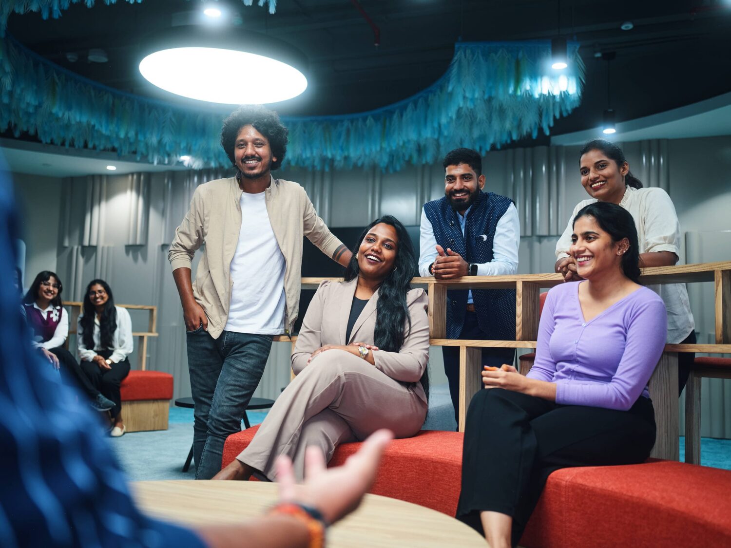 A group of casually dressed people in a modern lounge sit and stand around red seating under a circular ceiling light, smiling.