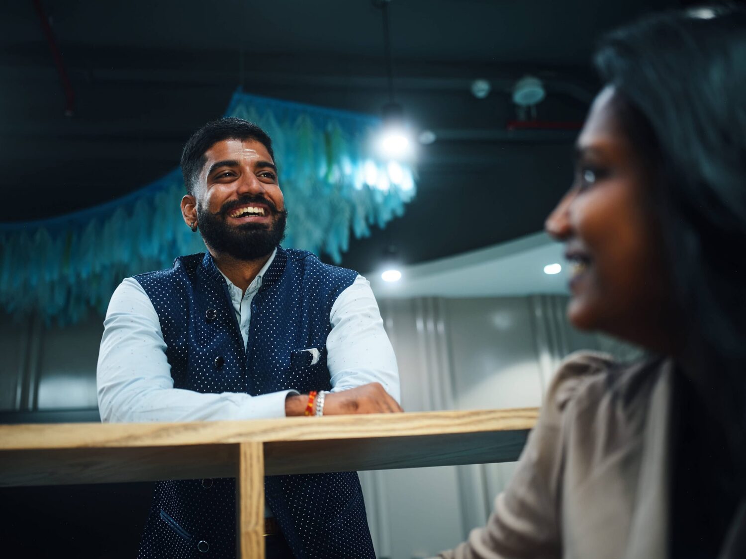 Smiling bearded man in a denim vest sits at a wooden table opposite a blurred woman in an office.
