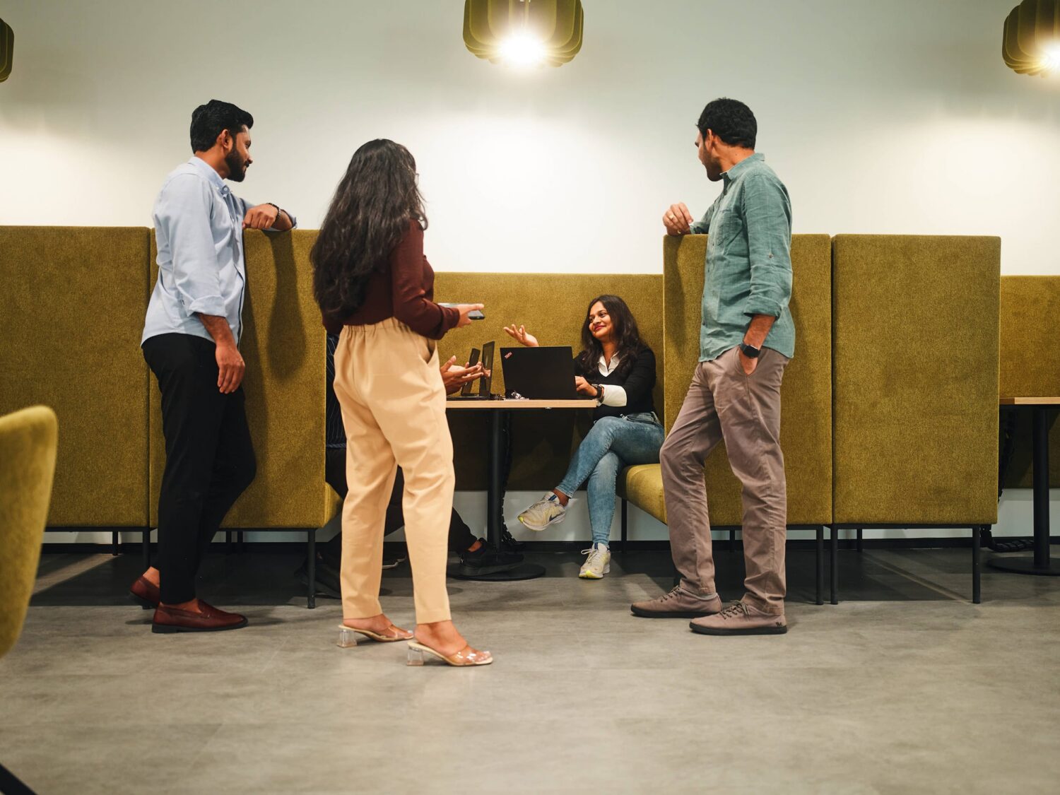 Four casually dressed people stand and chat in a modern cafe, with two others seated at a table in the background.