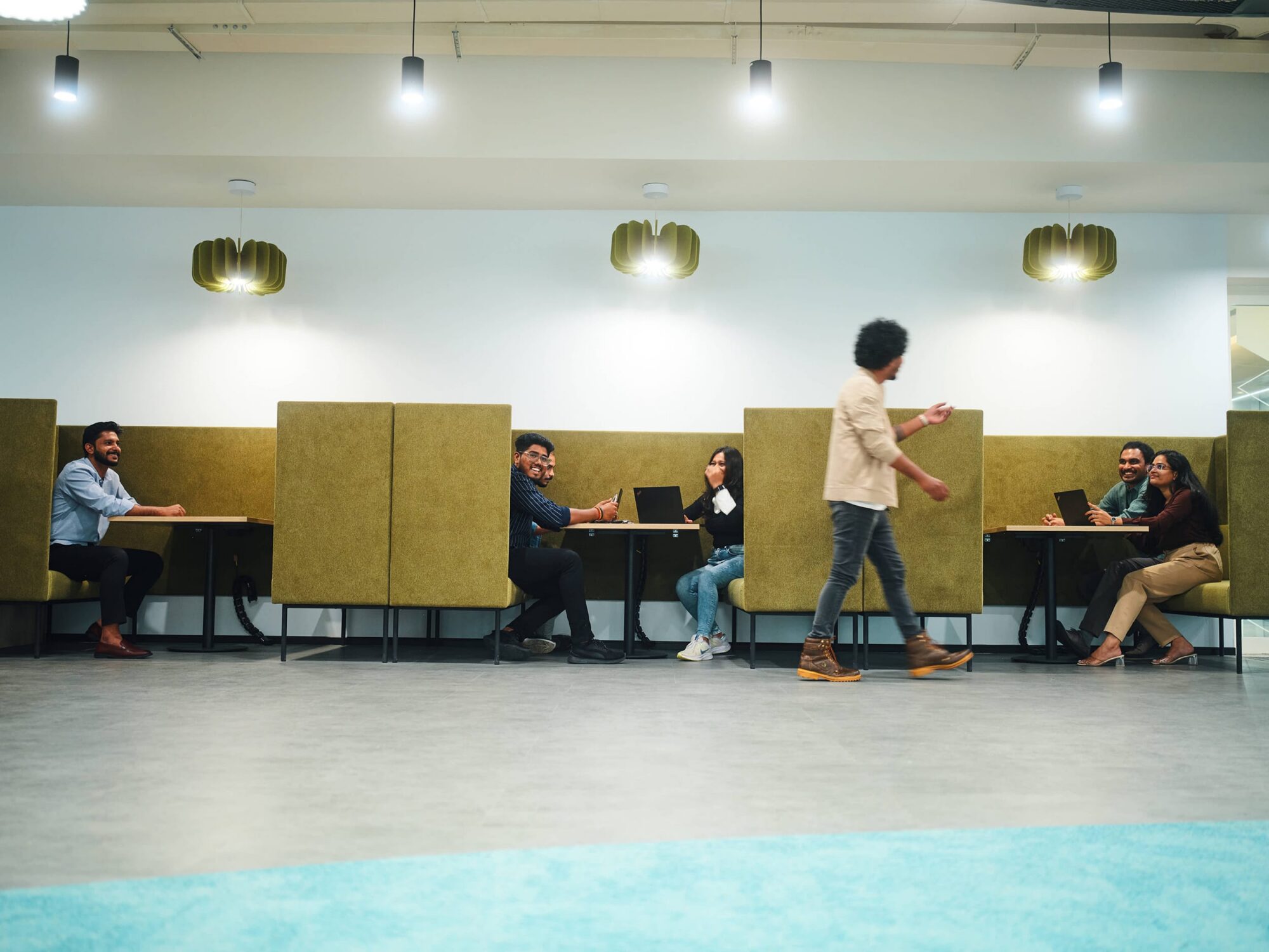 A person walks past cubicle partitions in a large, well-lit room where several people sit at tables behind the dividers.
