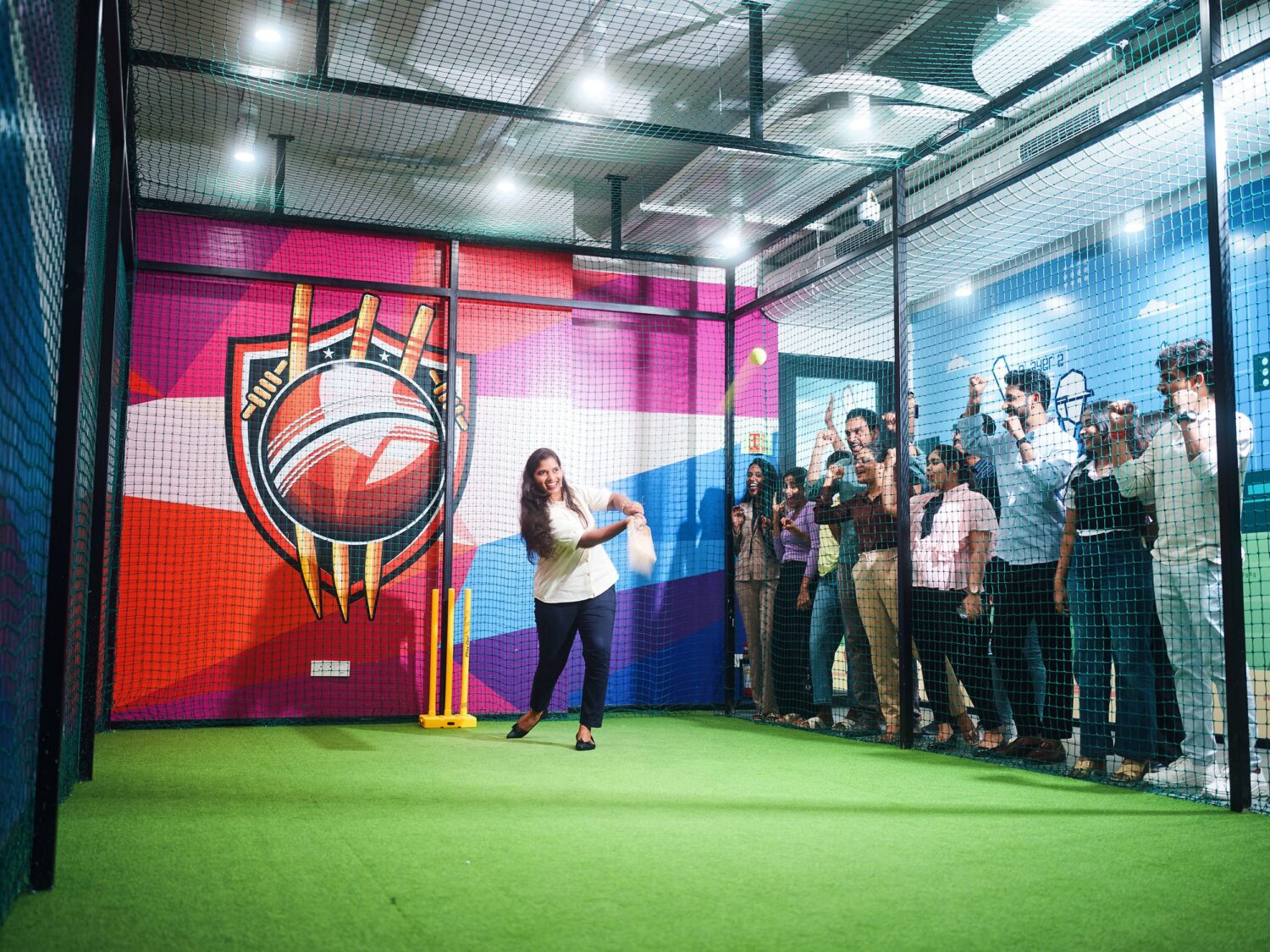 A woman in a white shirt addresses a group of people inside a colorful, netted indoor sports cage with a basketball mural and a green floor.