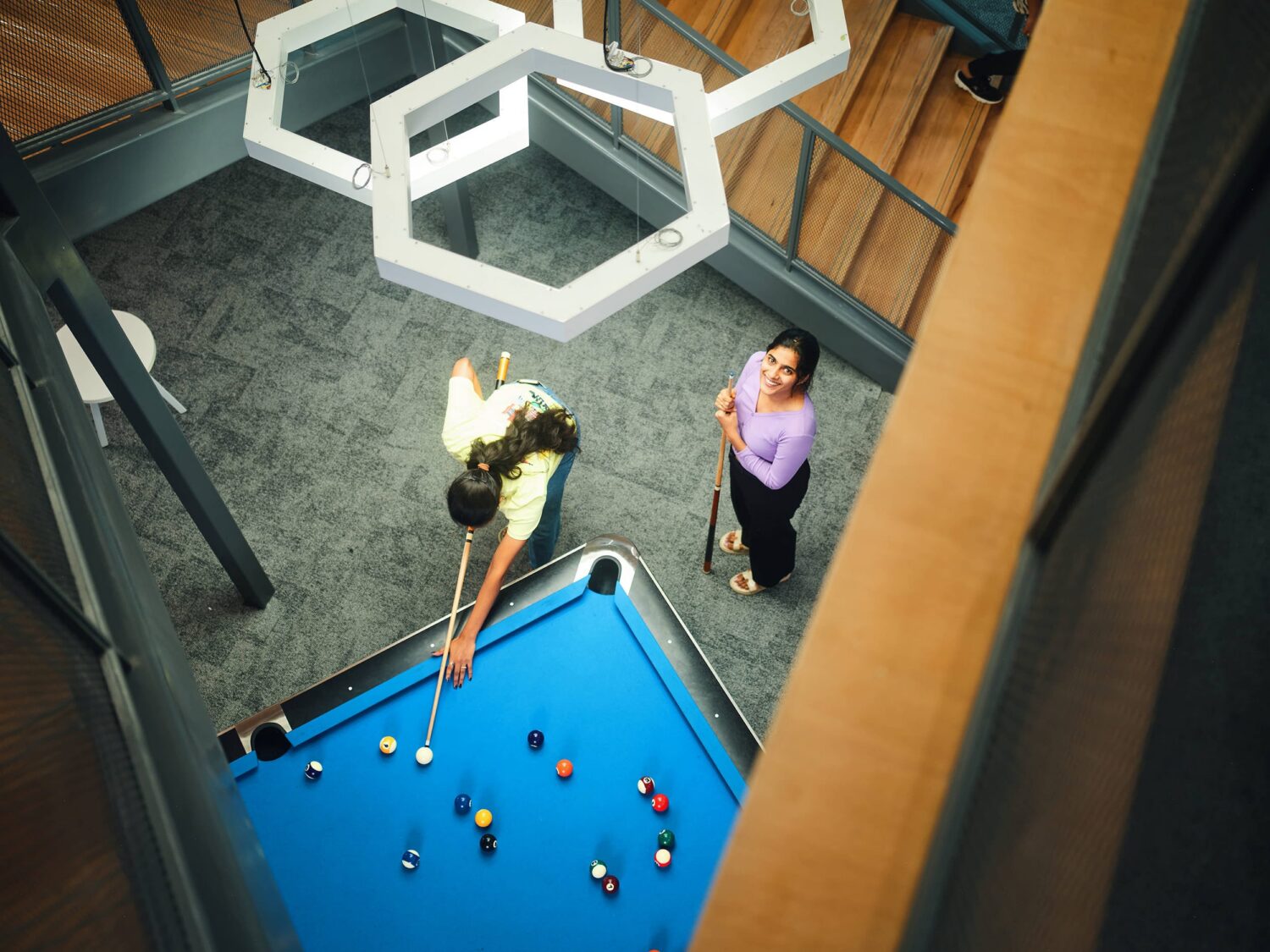 Two people stand around a blue pool table in a modern indoor space, viewed from above, beneath white hexagonal light fixtures.