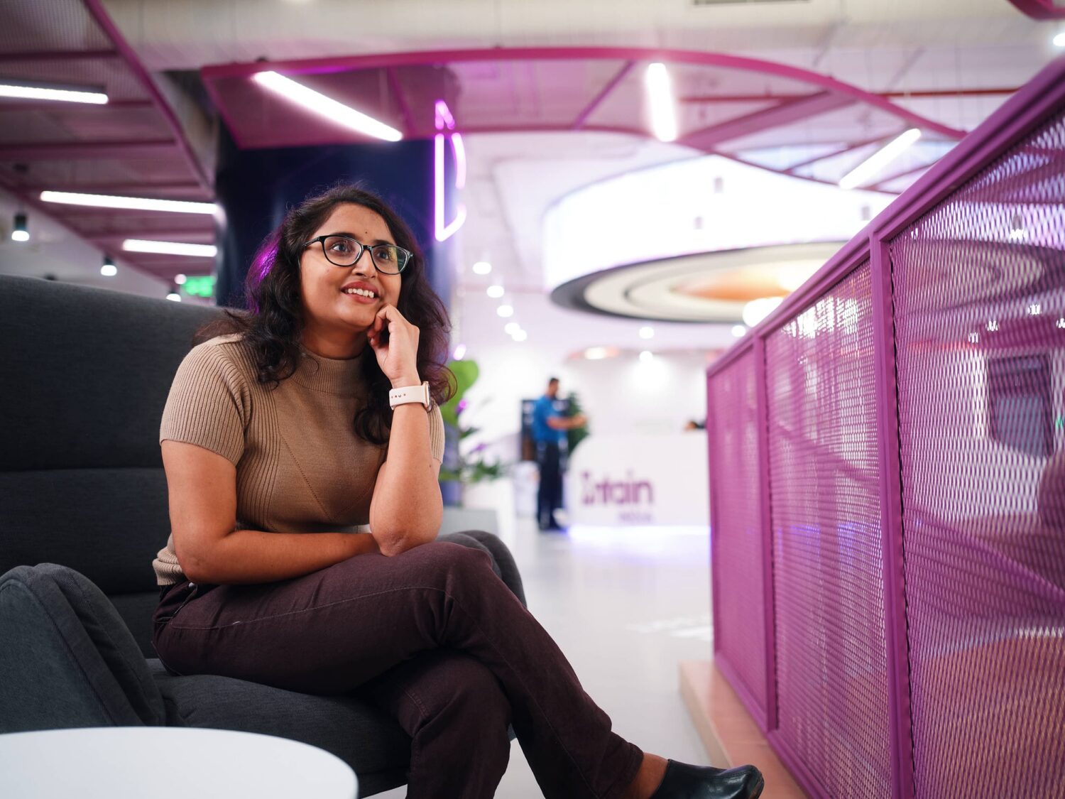 A smiling woman with glasses sits on a white chair in a bright, modern office space with purple railings.