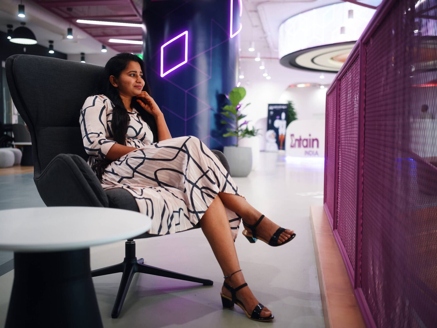 A woman in a white and black patterned dress sits cross-legged on a modern lounge chair in a stylish indoor space with purple lighting and a purple mesh divider.
