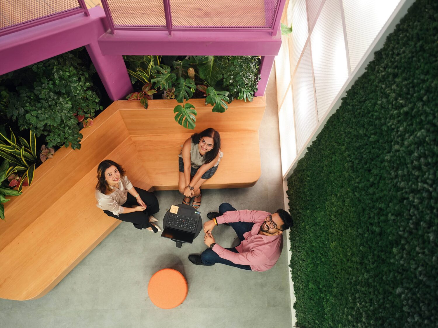 Top-down view of four people seated around a small orange round table in a plant-filled lounge with wooden benches.