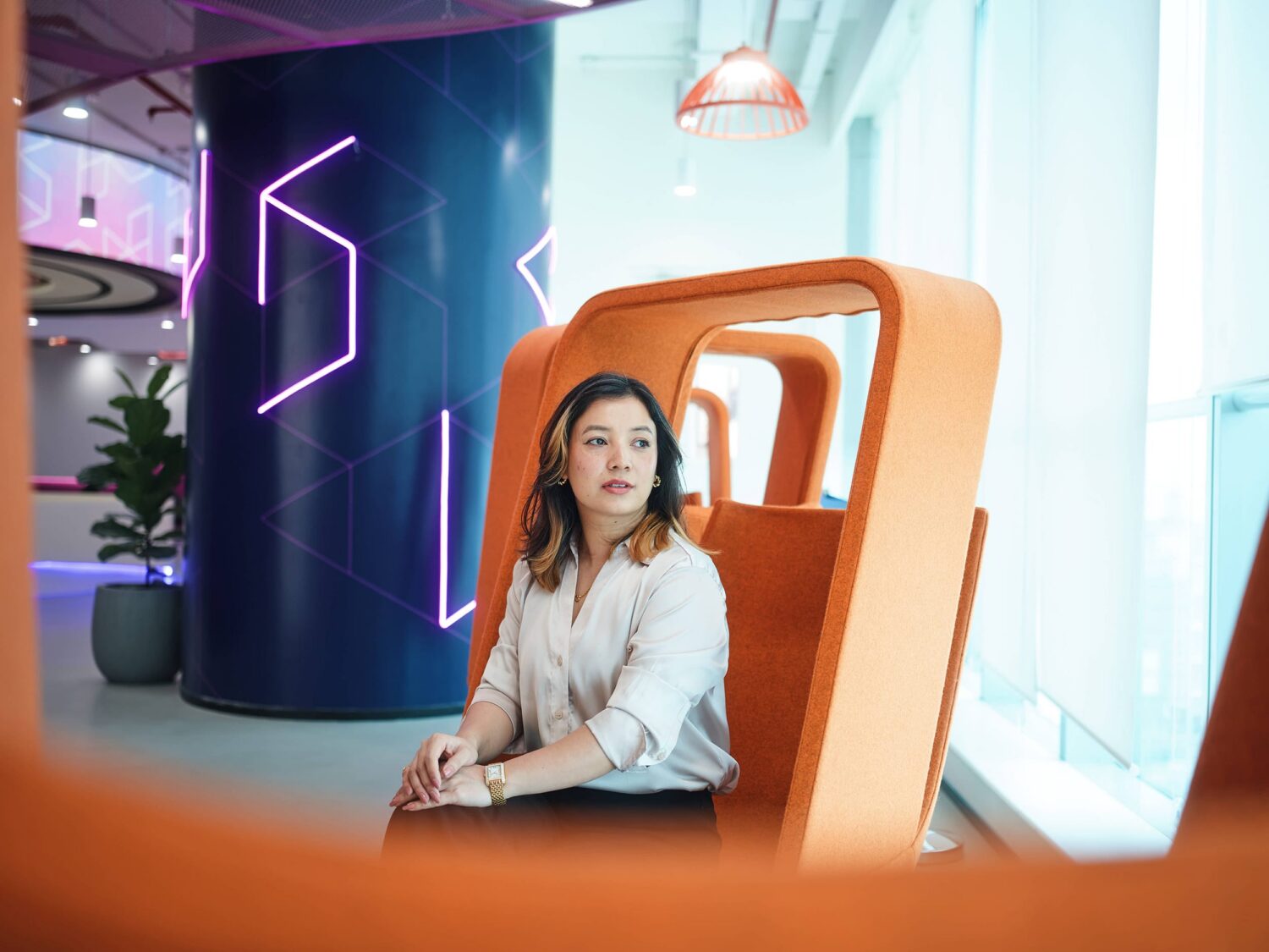 A woman sits in a large orange modern chair in a brightly lit, futuristic space with neon purple light accents.