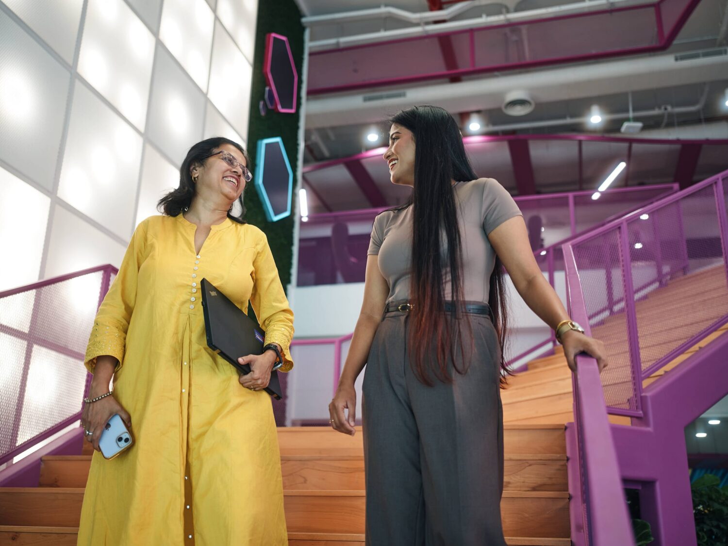 Two women stand on an indoor staircase with purple railings, smiling at each other; one in a yellow dress holding a notebook and phone, the other in gray overalls.
