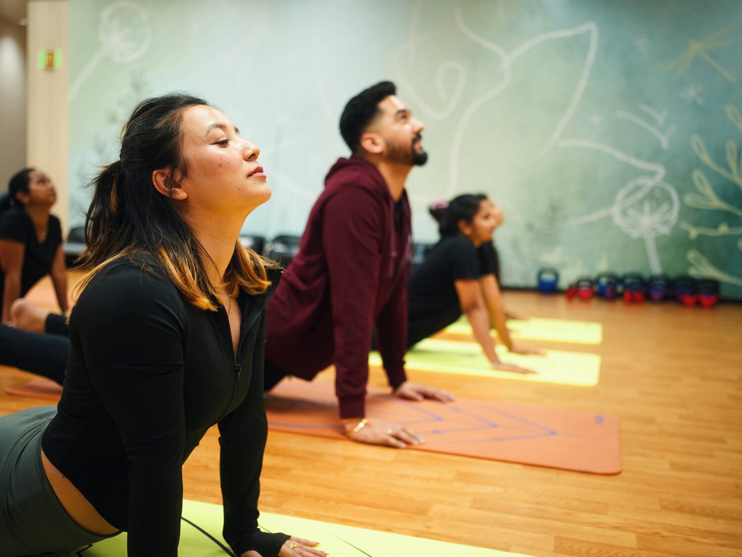 A group of people practice yoga on mats in a studio with a leafy mural on the wall.