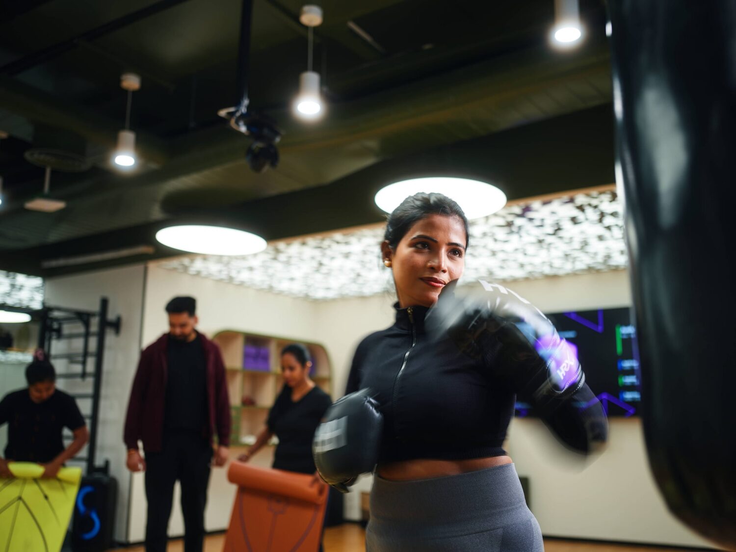 A woman wearing boxing gloves punches a bag in a gym, with other people watching in the background.