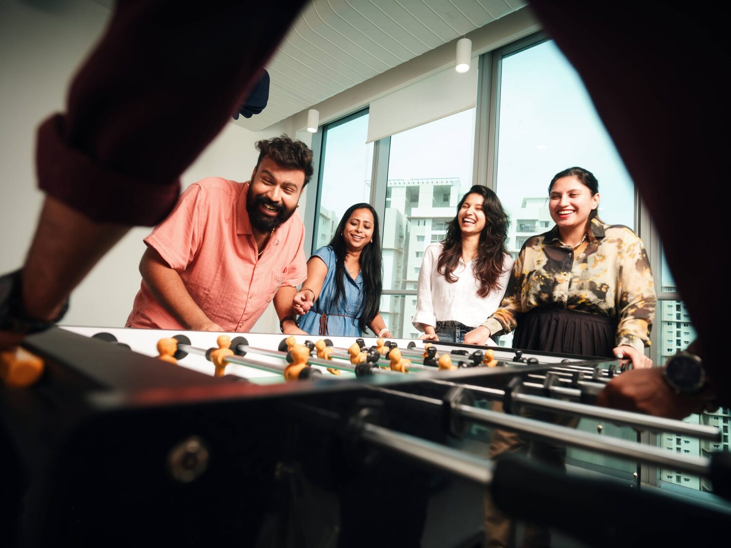Four friends gathered around a foosball table, smiling at the game in a bright room with large windows.