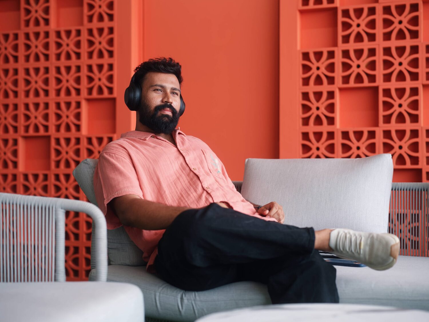 Bearded man in a peach shirt sits on a light gray outdoor sofa with a red lattice wall behind him.