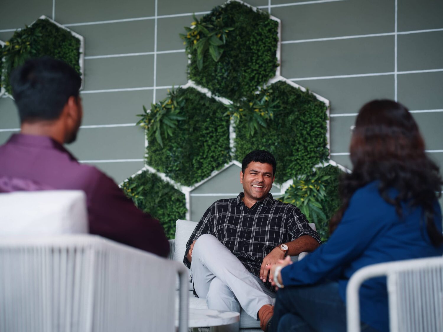 Three people sit in a studio, engaged in a discussion, with a geometric wall of green plants behind them.