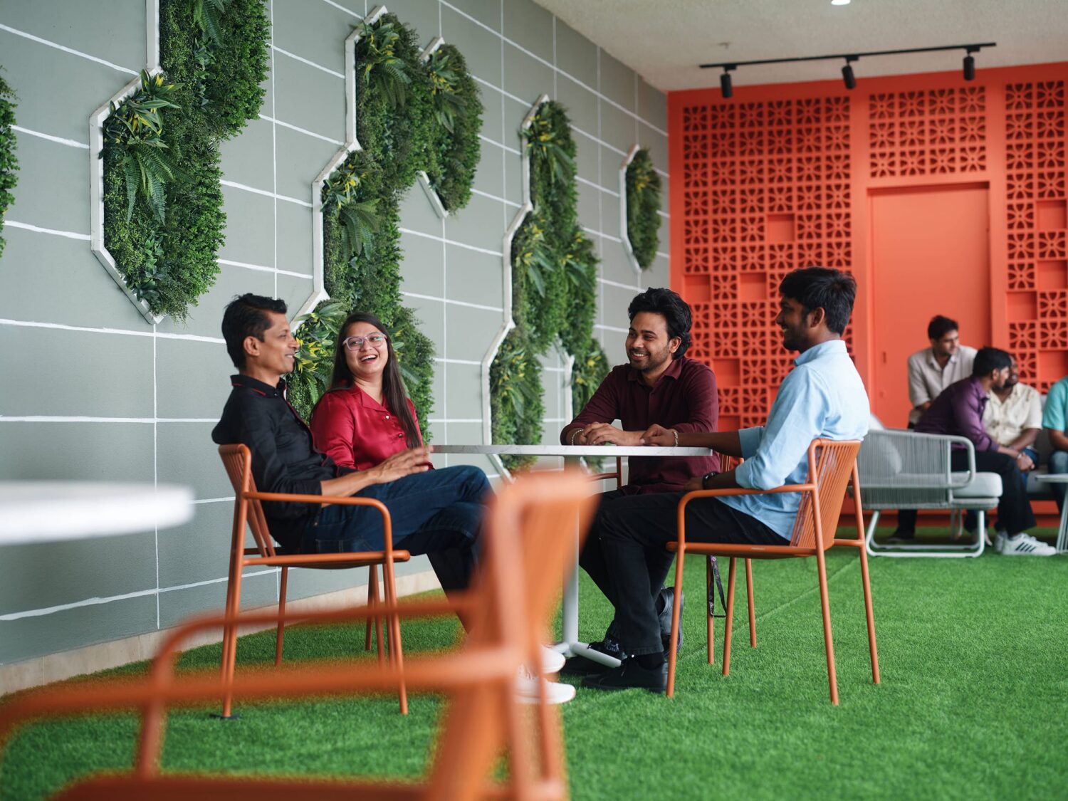 A group of people sits around a small table in a modern lounge with artificial grass flooring and greenery on the walls.