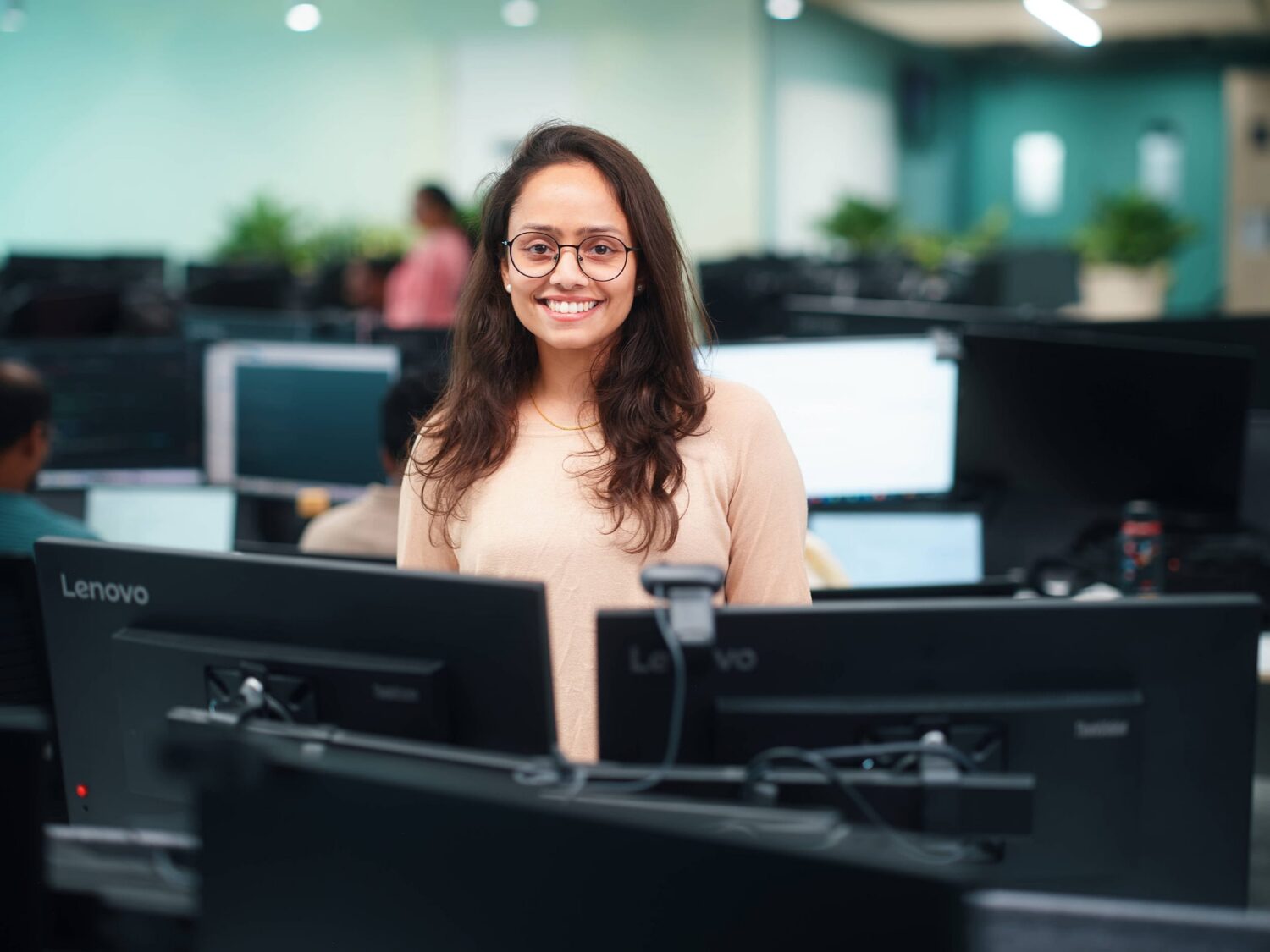 A smiling woman with glasses sits at a workstation in an office surrounded by multiple computer monitors.