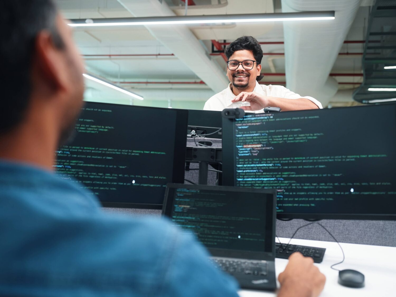 Two programmers work at a desk with multiple monitors displaying code; one wearing a blue shirt sits facing away while the other stands nearby, smiling.
