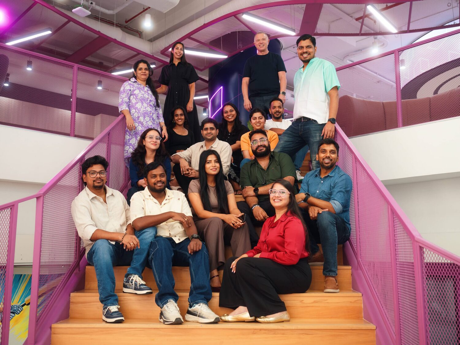 A diverse group of people poses on a wooden staircase inside a bright, pink-accented indoor space.