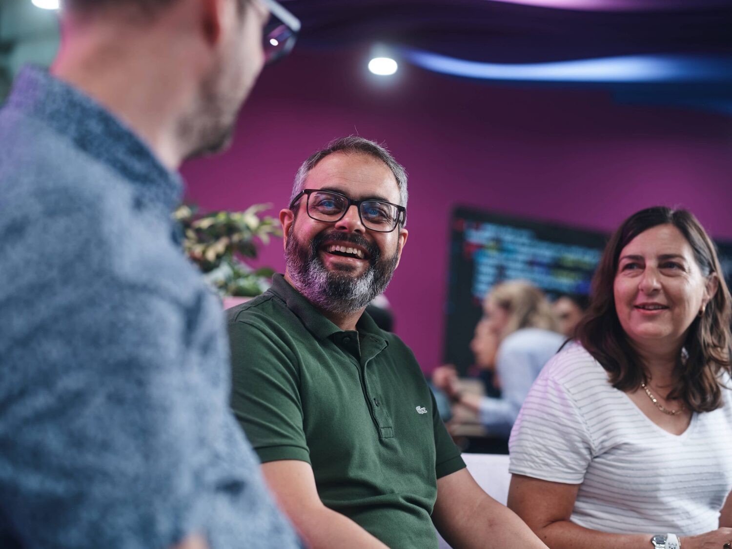 Bearded man in a green polo smiles at the man in the foreground while a woman in a white shirt watches in a colorful indoor setting.