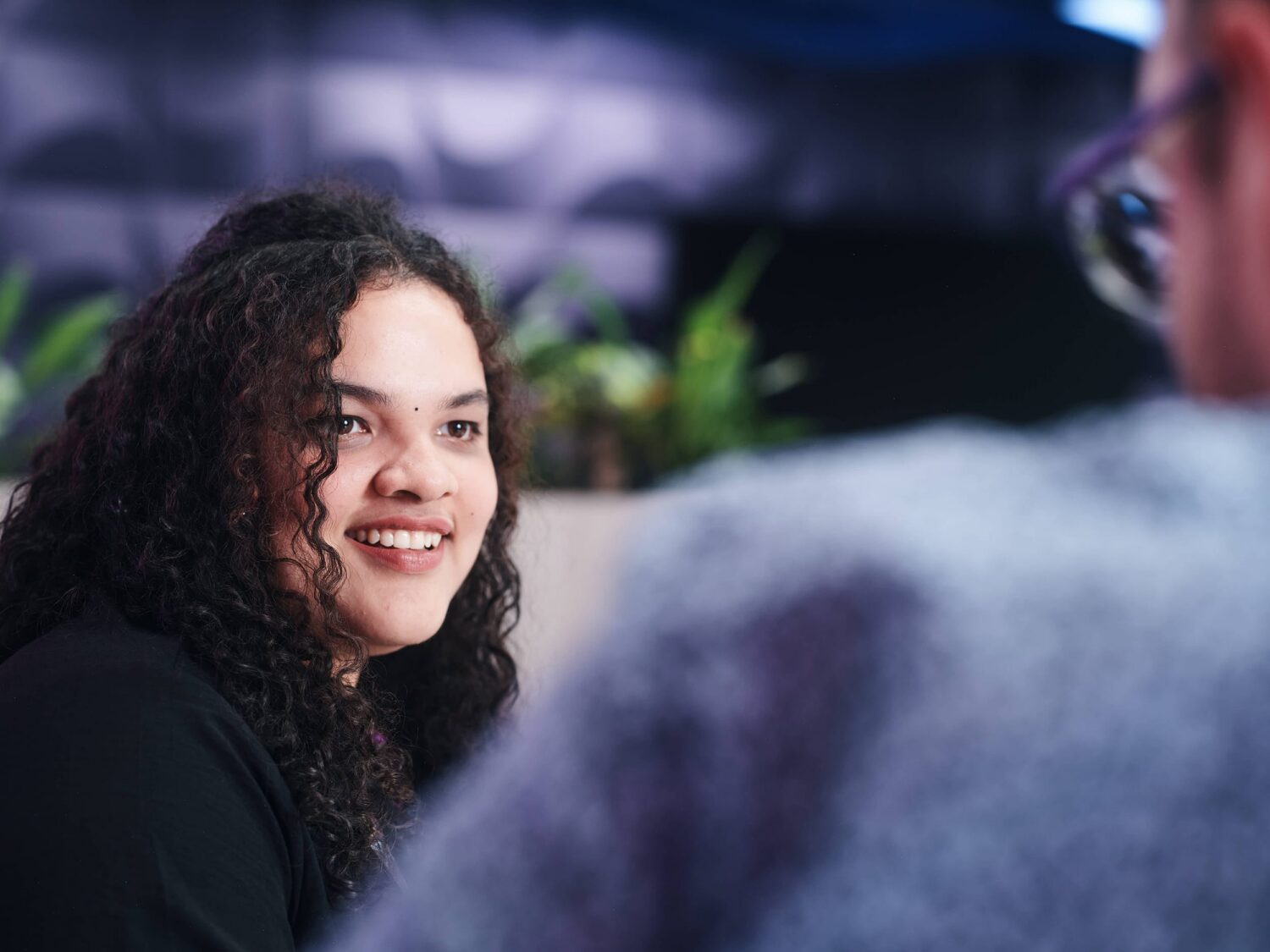 A woman with curly dark hair wearing a black top smiles while talking to someone in the foreground.