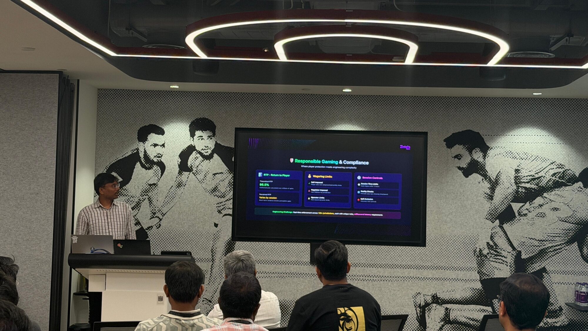 A presenter speaks at a podium in a conference room while attendees watch a slide on a screen, with a black-and-white mural of cricketers on the wall.