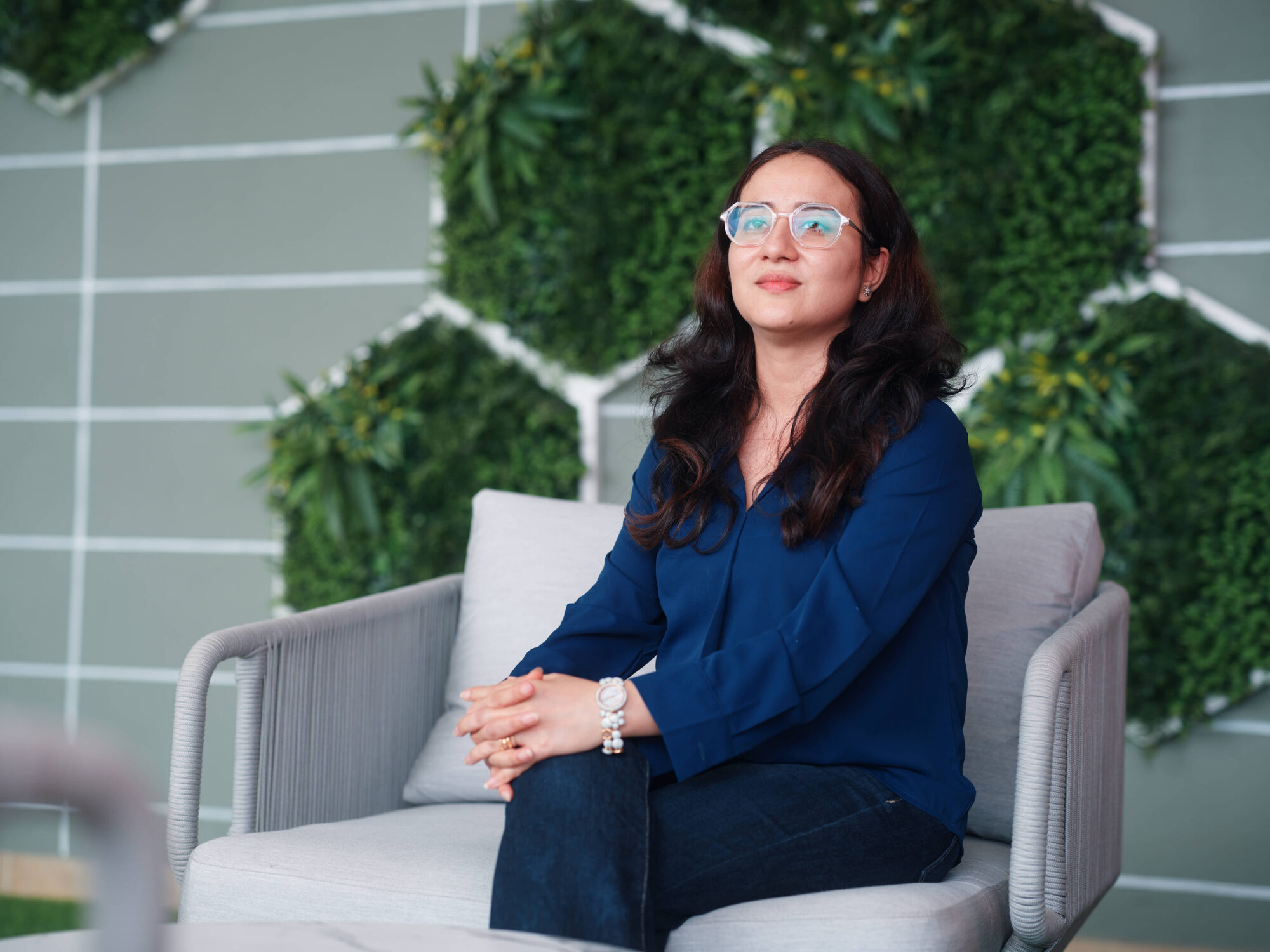 Woman with dark hair and glasses sits on a light chair in a modern room with a green plant wall behind her.